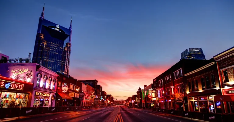 Broadway in downtown Nashville at sunrise with neon signs and the AT&T Building in the background.