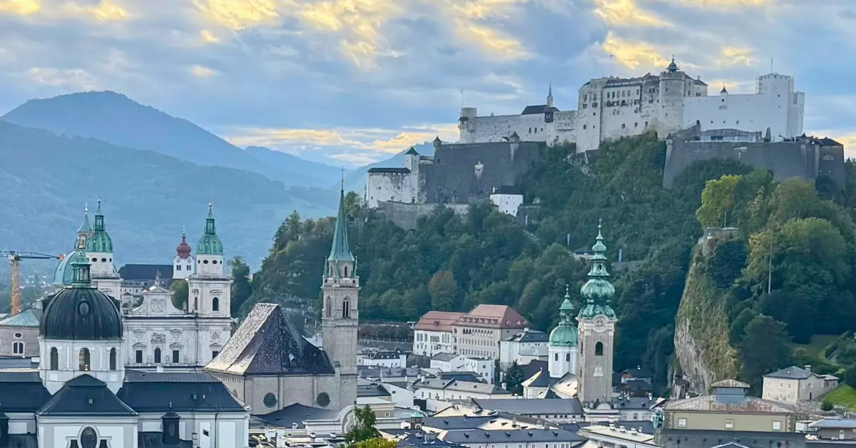 Hohensalzburg Fortress overlooking Salzburg's historic Old Town and church towers, with mountains and a dramatic cloudy sky in the background.