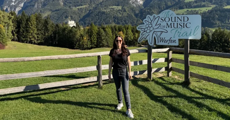 Standing beside the Sound of Music Trail sign at the Sound of Music Meadow in Werfen, Austria, with the Austrian Alps in the background.