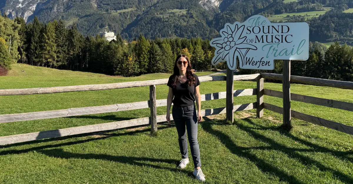 Standing beside the Sound of Music Trail sign at the Sound of Music Meadow in Werfen, Austria, with the Austrian Alps in the background.