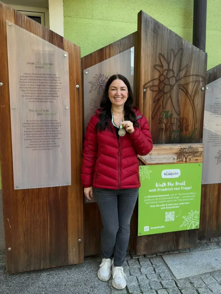 Holding up a Sound of Music Trail completion medal while standing in front of the trail information signs in Werfen, Austria.