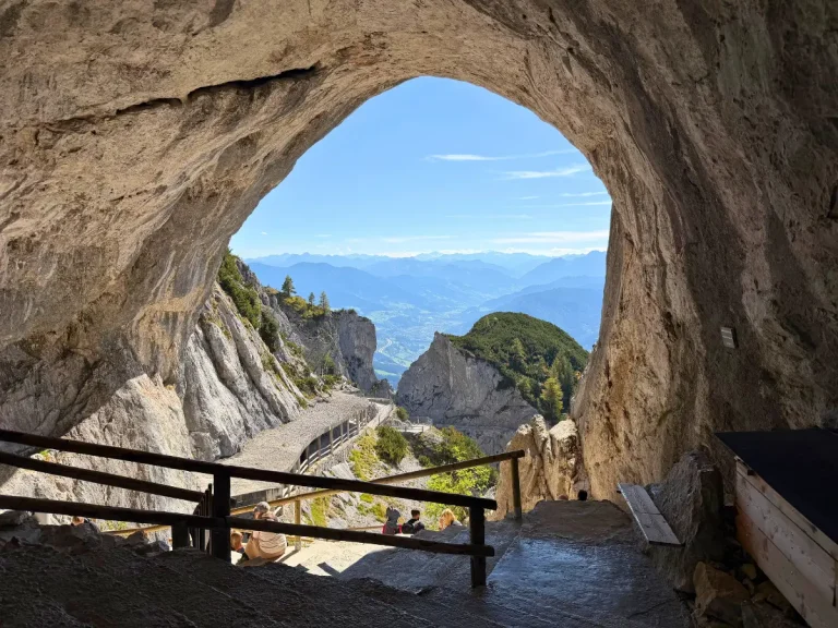 View of the Austrian Alps through the rocky entrance of Eisriesenwelt Ice Cave, with a walking path winding down the mountain.
