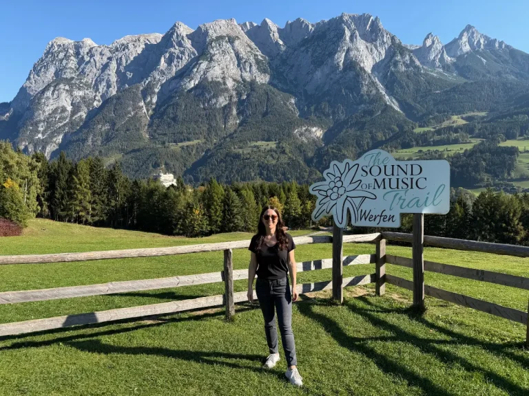Traveler standing beside the Sound of Music Trail sign in Werfen, Austria, with a green meadow and the Austrian Alps and Burg Hohenwerfen in the background.